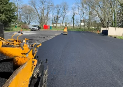 Fresh asphalt being paved and rolled on a large lot in Toms River, with trees and vehicles in the background—showcasing expert asphalt services for any project size.