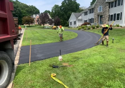 Workers from Asphalt Worxx LLC paving a curved asphalt driveway in front of suburban houses on a sunny day.