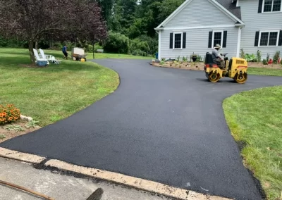 Workers paving and smoothing fresh asphalt on a residential driveway beside a gray house with a garden, showcasing quality asphalt services in New Jersey.