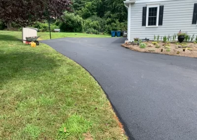 Freshly paved black asphalt curves past grass and a white house with a garden bed—expertly finished by Asphalt Worxx LLC, your trusted choice for asphalt services.
