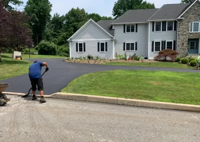 A person from Asphalt Worxx LLC smooths newly paved asphalt on a driveway in front of a suburban house, showcasing expert asphalt services.
