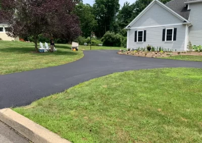 A freshly paved asphalt driveway by Asphalt Worxx LLC curves toward a gray house in Toms River, surrounded by green lawns and trees.