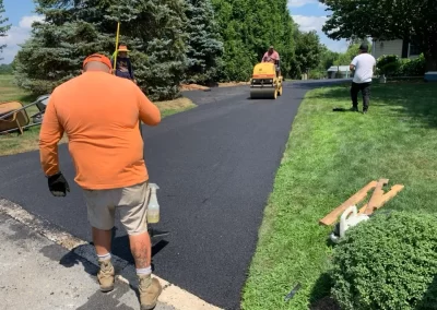 Three workers provide asphalt services in Toms River, paving a driveway with fresh asphalt—one uses a roller while two handle tools on a sunny day.