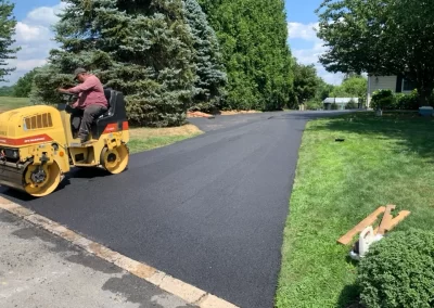 A worker from Asphalt Worxx LLC drives a yellow steamroller to flatten freshly laid asphalt on a driveway next to green grass and trees, showcasing expert asphalt services in New Jersey.