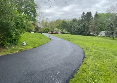 Freshly paved asphalt driveway curving through a green lawn, with trees and parked cars in the distance—showcasing quality asphalt services in New Jersey.