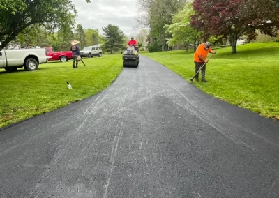 Three workers from Asphalt Worxx LLC pave and smooth a fresh black asphalt driveway in Toms River, with green grass and parked trucks nearby, showcasing quality asphalt services.