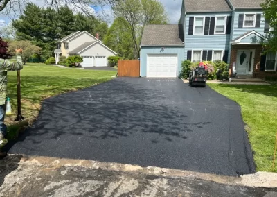 Freshly paved asphalt driveway in front of a blue house in Toms River, New Jersey, with a worker from Asphalt Worxx LLC standing to the left side.