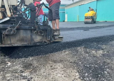 Workers operate paving equipment to lay fresh asphalt on a road in Toms River, New Jersey, with a roller compacting in the background.
