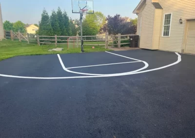 A driveway basketball court in Toms River with freshly painted, uneven white lines sits beside a house and fenced yard, highlighting recent asphalt services.