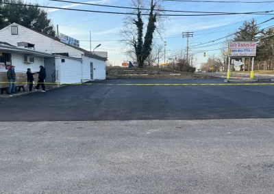 Freshly paved asphalt parking lot cordoned off with caution tape; a few people stand near a white building on the left, highlighting quality asphalt services in Toms River.