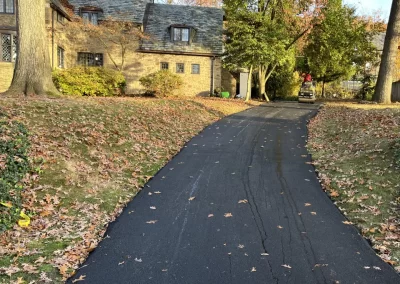 A paved driveway leads to a brick house surrounded by autumn trees and fallen leaves, showcasing quality asphalt services in Toms River.
