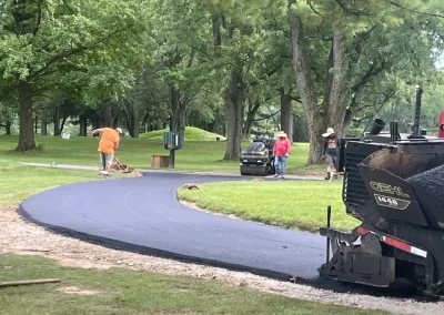 Workers from Asphalt Worxx LLC pave a winding path with fresh asphalt in a green, tree-filled park in New Jersey.