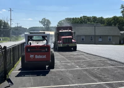 A Bobcat and a dump truck from Asphalt Worxx LLC are working in a sunny, mostly empty parking lot near a building, preparing the site for premium asphalt services.