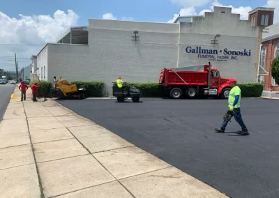 Workers from Asphalt Worxx LLC are repaving a parking lot near a funeral home in New Jersey on a sunny day, with paving equipment and a red truck on site providing expert asphalt services.