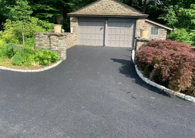 Freshly paved black asphalt driveway by Asphalt Worxx LLC leads to a stone garage, surrounded by green plants and shrubs.