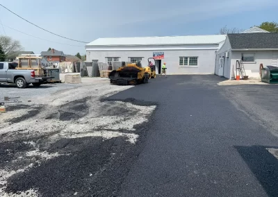Workers pave an asphalt driveway in front of a white commercial building under a clear sky in Toms River, New Jersey.