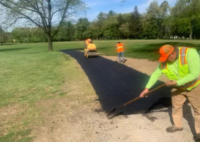 Three workers in safety vests pave a new asphalt path through a grassy New Jersey park on a sunny day, showcasing expert asphalt services.
