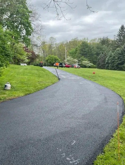 A newly paved asphalt driveway curves through green grass toward workers and vehicles in the distance.